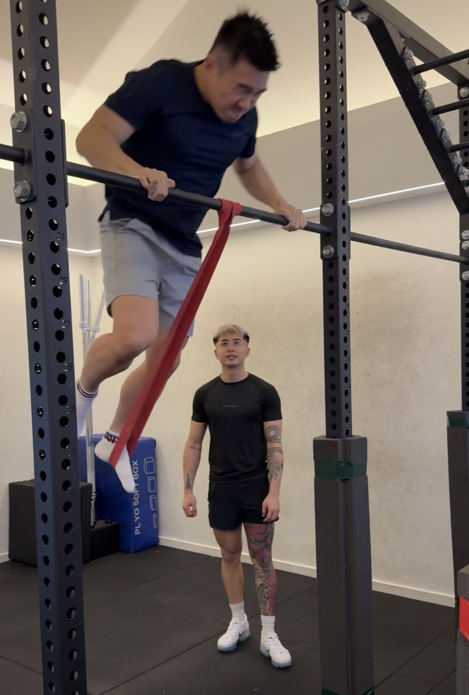 A client attempting a banded muscle-up on the bar while Fred supervises from behind.